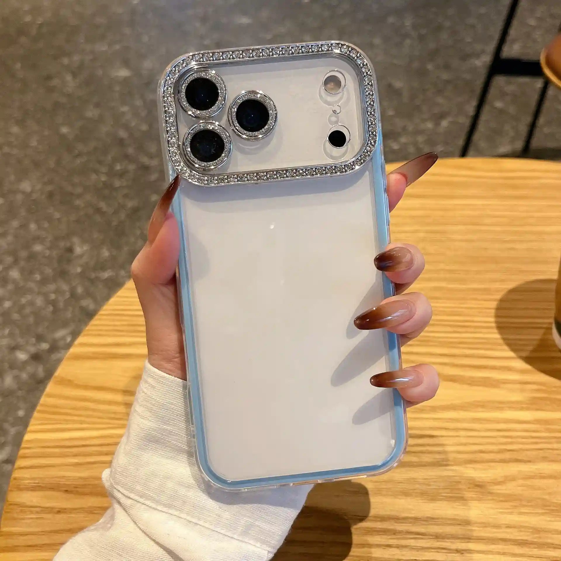 A minimalist light blue hybrid phone case featuring a glamorous silver rhinestone-embedded camera lens protection frame and a crystal clear transparent back, held in hand.