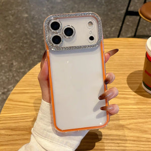 A minimalist vibrant orange hybrid phone case featuring a clear back panel and a glittering diamond rhinestone protective ring around the camera lenses.