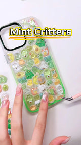 Handcrafted phone case with a mint green frame, densely filled with a 3D collage of glossy, transparent green frog charms, cartoon dinosaurs, split avocados, and golden crescent moons.