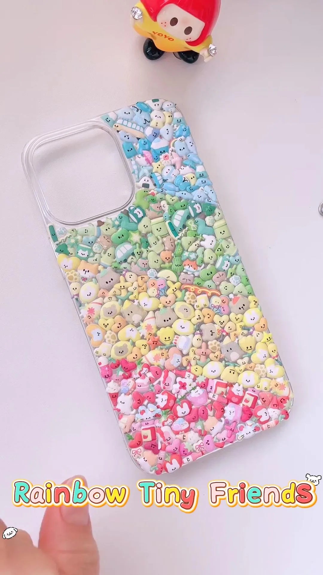 Handcrafted crystal clear phone case densely packed with a 4-tier rainbow gradient collage of tiny glossy cartoon charms transitioning from sky blue to green, yellow, and vibrant red.