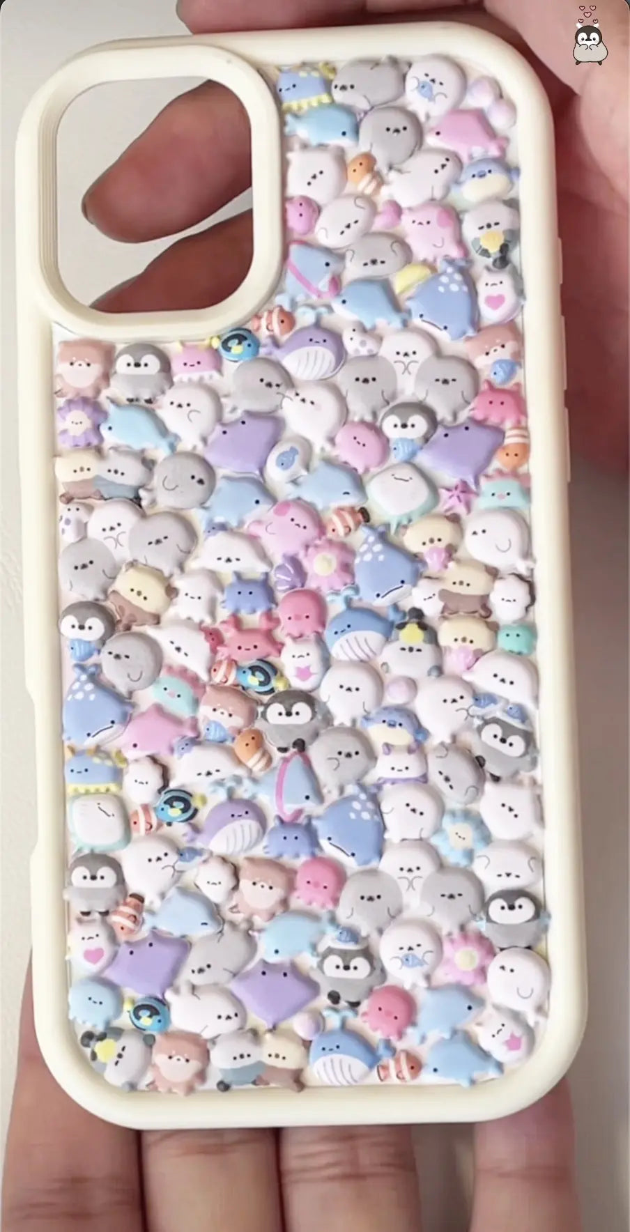 Finished DIY iPhone case with cream frame, decorated with dense 3D cute sea creature stickers, including seals, penguins, and other kawaii marine animals in pastel colors.

