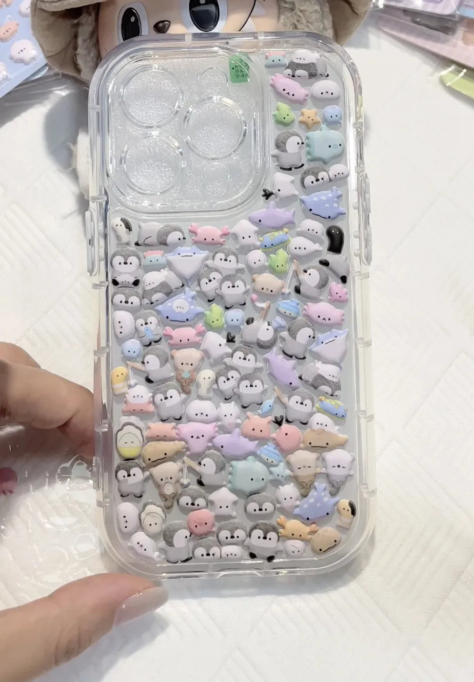 Finished DIY clear iPhone case decorated with dense mini positive energy penguin and sea animal stickers, featuring a soft pastel kawaii aesthetic.