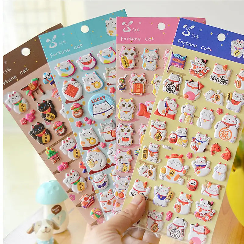 Display of multi-color "Fortune Cat" 3D puffy sticker set, showing cute lucky cat designs in light blue, pink, blue, and brown themes.