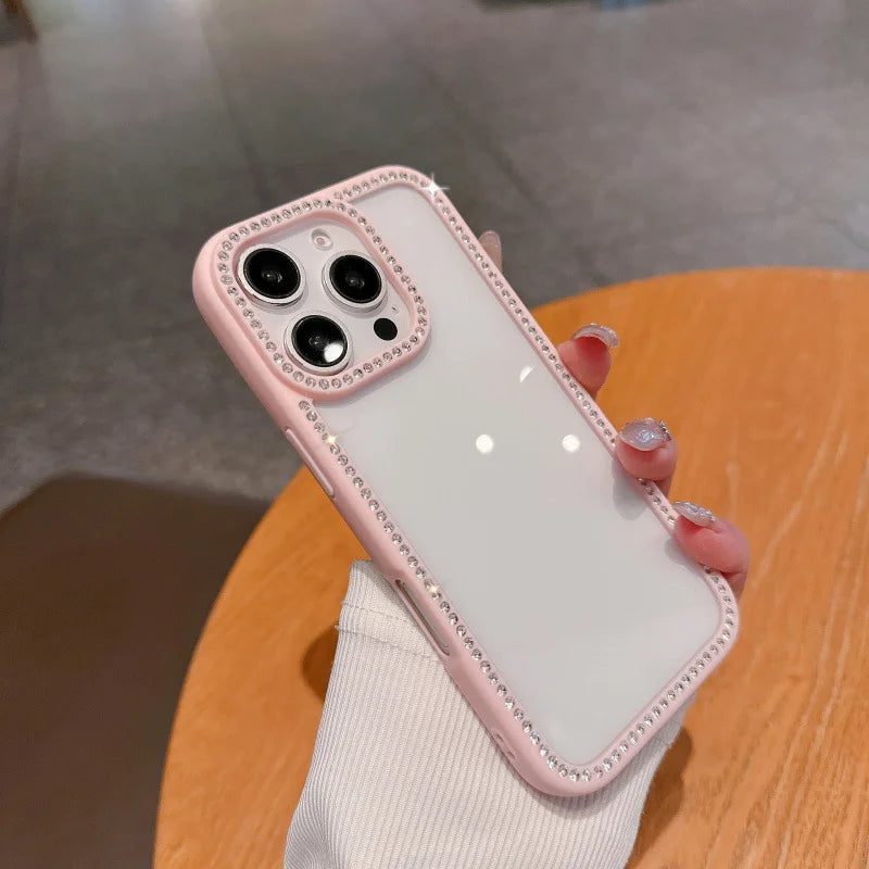 A luxury pastel pink bumper phone case with a clear back, featuring a full frame of glittery rhinestones around the border and camera lenses, held in hand.