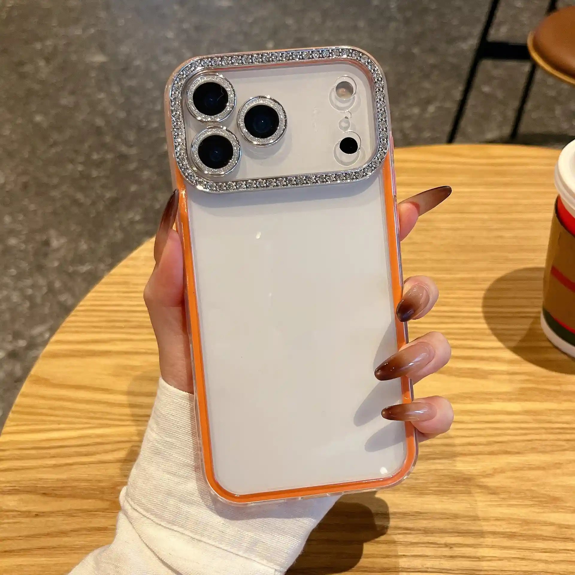 A minimalist vibrant orange hybrid phone case featuring a clear back panel and a glittering diamond rhinestone protective ring around the camera lenses.